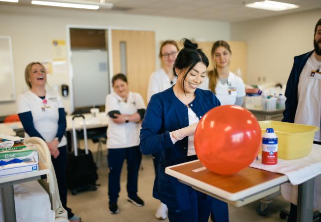 A person in a nursing classroom practices with a balloon while others in white coats watch. 