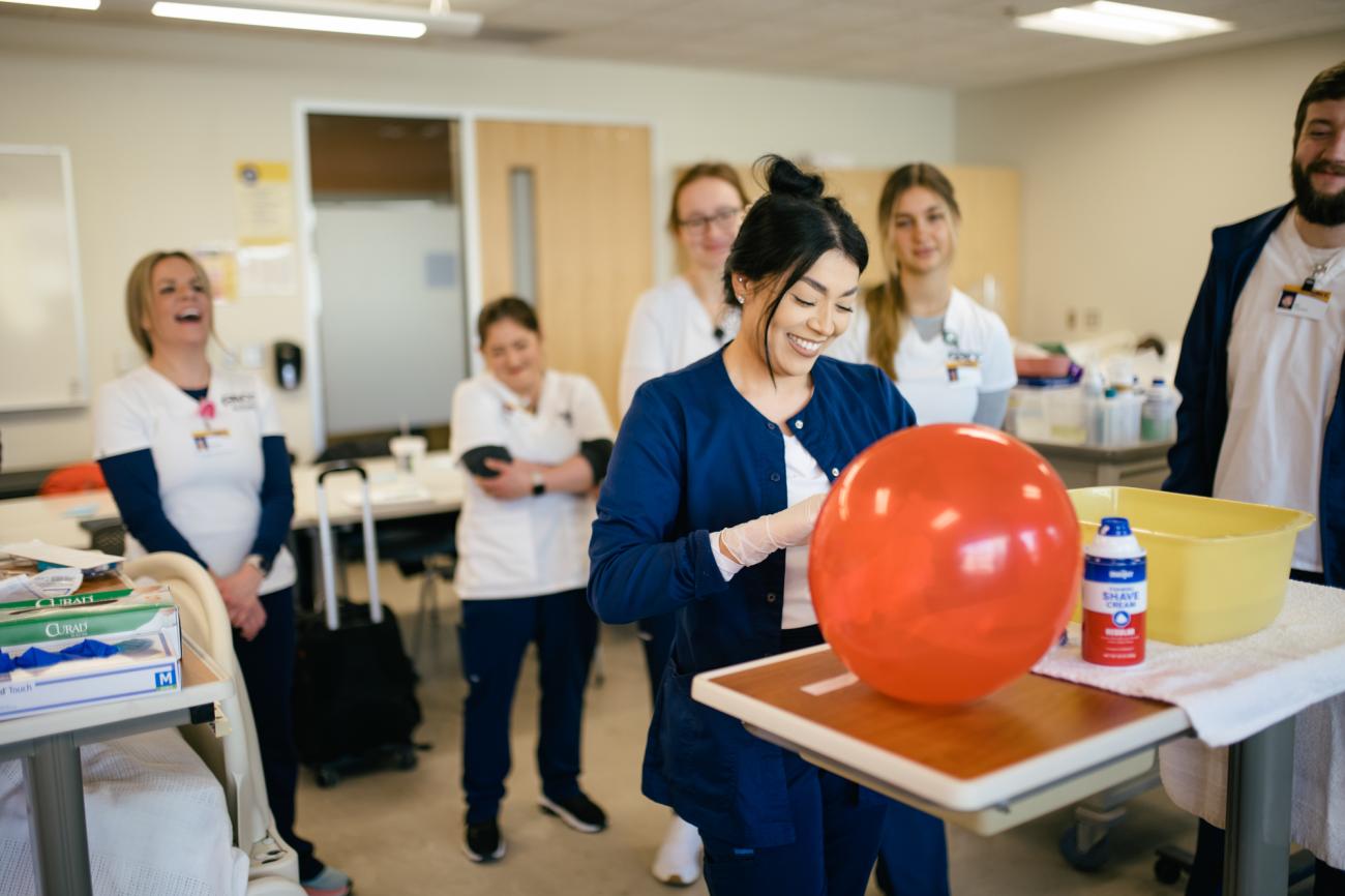 A person in a nursing classroom practices with a balloon while others in white coats watch. 