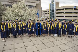 Dr. Lepper poses with a large group of students, all wearing caps and gowns and a yellow stole. 