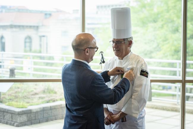 GRCC professor Gilles Renusson, wearing a chef's coat and hat, is presented with the French medal of Chevalier du Merite Agricole from Consul General Guillaume Lacroix.