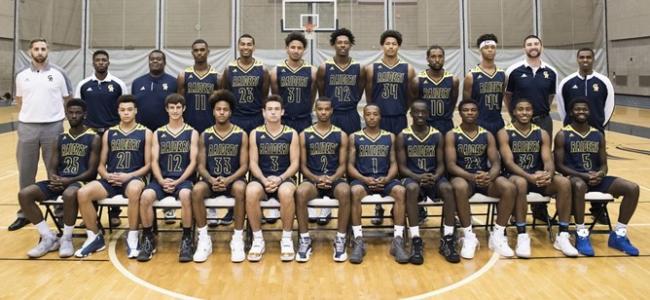 The members of the 2017-18 men's basketball team are in two rows, with the first row seated, on the fieldhouse floor.