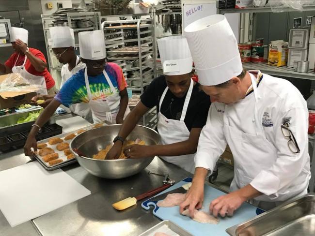Chef Bob Schultz shows two high school students how to bread fish.