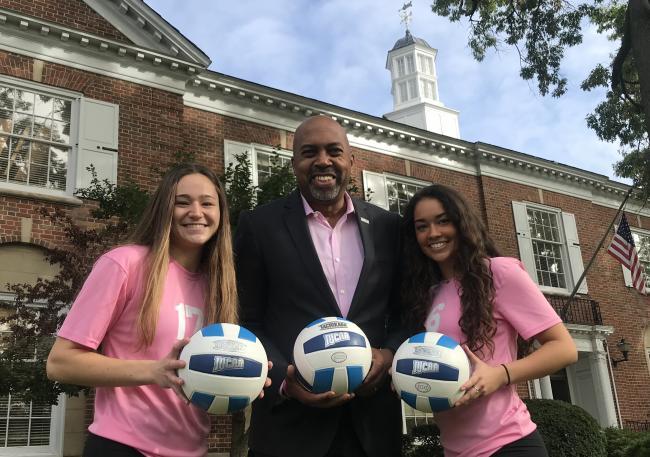 Volleyball players Karissa Ferry and Audrey Torres surround President Bill Pink, wearing pink uniforms.