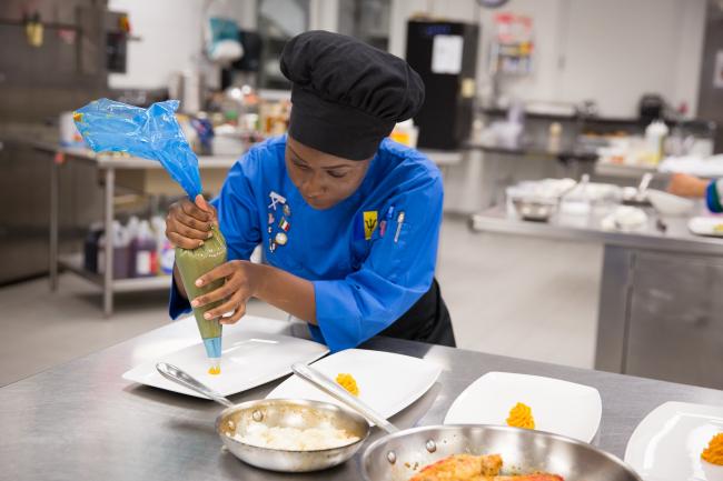 Student from Barbados in a black chef's hat and blue kitchen coat squeezing ingredients from a funnel during competition.