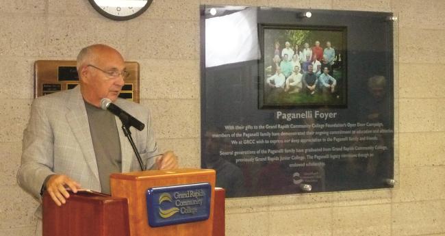 Carl Paganelli Sr. Standing behind a GRCC podium at the dedication of the Paganelli Family Foyer in 2013. A large plaque with a photo of his family is on the wall behind him.