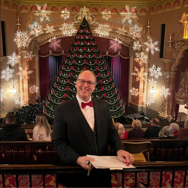 GRCC Music program alumnus Shawn Lawton poses in a suit and bowtie in front of a Christmas tree.