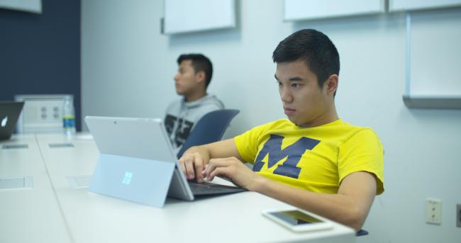 Student wearing a yellow University of Michigan t-shirt working with a laptop computer.