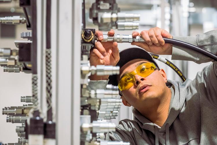 A male student wearing safety goggles works on manufacturing equipment.
