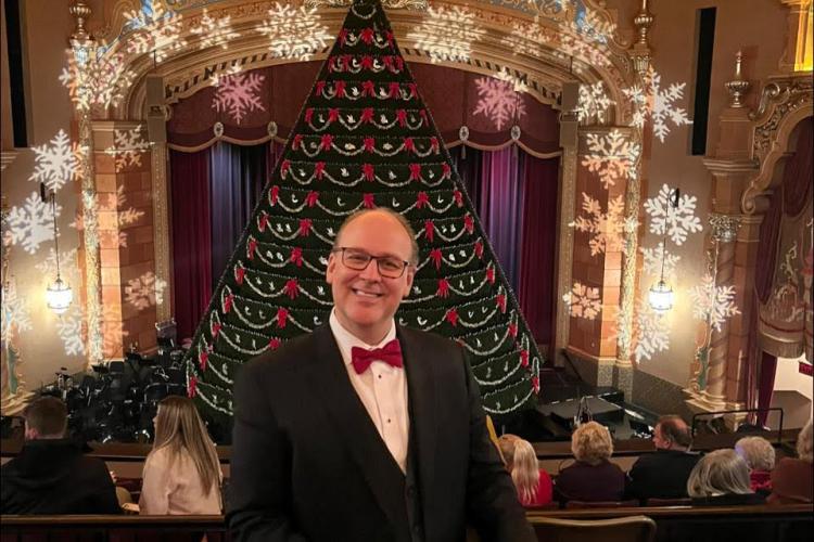 GRCC Music program alumnus Shawn Lawton poses in a suit and bowtie in front of a Christmas tree.