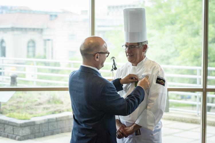 GRCC professor Gilles Renusson, wearing a chef's coat and hat, is presented with the French medal of Chevalier du Merite Agricole from Consul General Guillaume Lacroix.