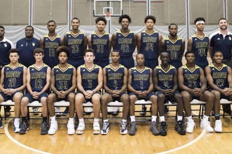 The members of the 2017-18 men's basketball team are in two rows, with the first row seated, on the fieldhouse floor.