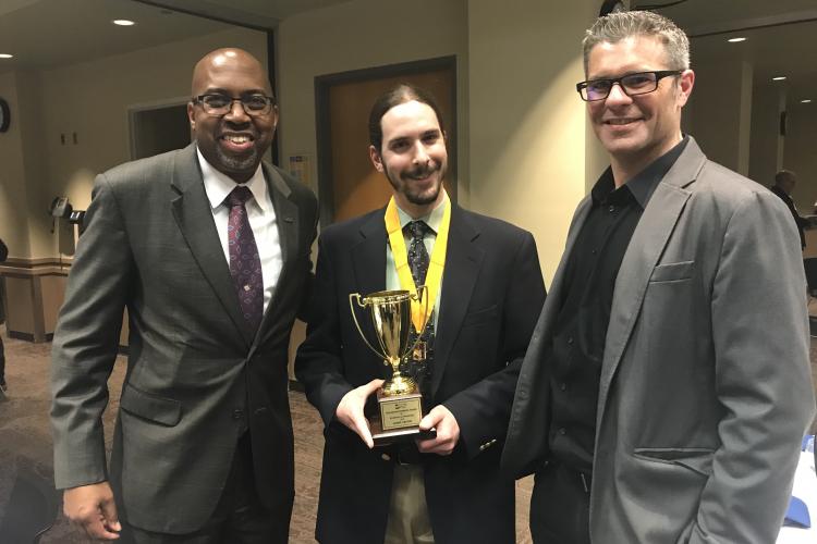 GRCC Andrews Award winner Daniel Cruzan wearing his gold medal and holding his award. GRCC President Bill Pink is on Daniel's right, and professor Andrew Rozema is on his left.