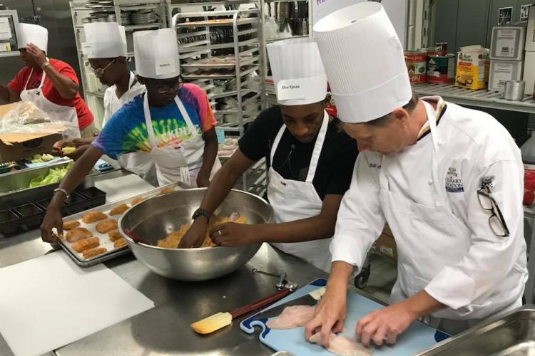 Chef Bob Schultz shows two high school students how to bread fish.