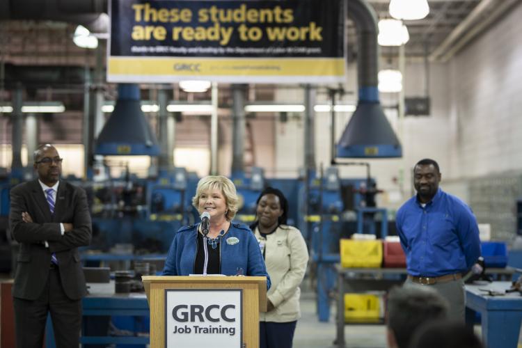 GRCC press conference participatants stand near a podium in the welding area of the Tassell-MTEC. Left to right: GRCC President Bill Pink; Dr. Amy Mansfield, dean of the school of workforce development; Elizabeth Pena, student in the medical assistant apprenticeship program, and Carlos Piggee, high-performance team coordinator at Johnson Controls