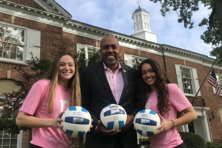 Volleyball players Karissa Ferry and Audrey Torres surround President Bill Pink, wearing pink uniforms.