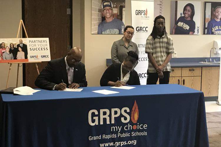 GRCC President Bill Pink and GRPS Superintendent Teresa Weatherall Neal seated at a table signing two documents that exted partnerships for concurrent enrollment and the Learning Center program. Students Najee Smith and Fatemeh Abedi are standing behind them. 