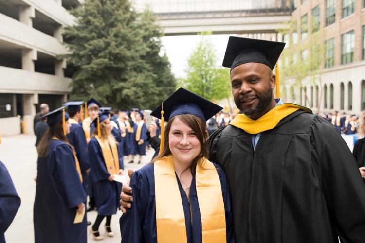 Two students in graduation caps and gowns with other graduates behind them.