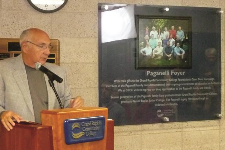 Carl Paganelli Sr. Standing behind a GRCC podium at the dedication of the Paganelli Family Foyer in 2013. A large plaque with a photo of his family is on the wall behind him.