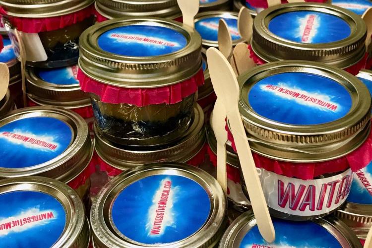 Small pies in a jar with "Waitress" written on the sides and the production website written on the lid. Each glass container has a wooden spoot attached.