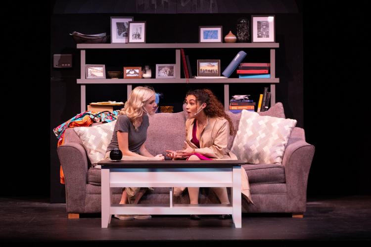 Performers rehearsing a scene on a set with a sofa and bookshelf.