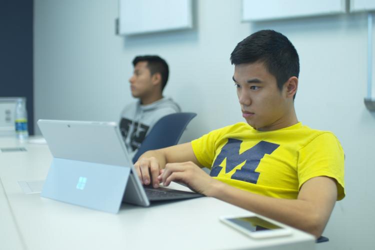 Student wearing a yellow University of Michigan t-shirt working with a laptop computer.