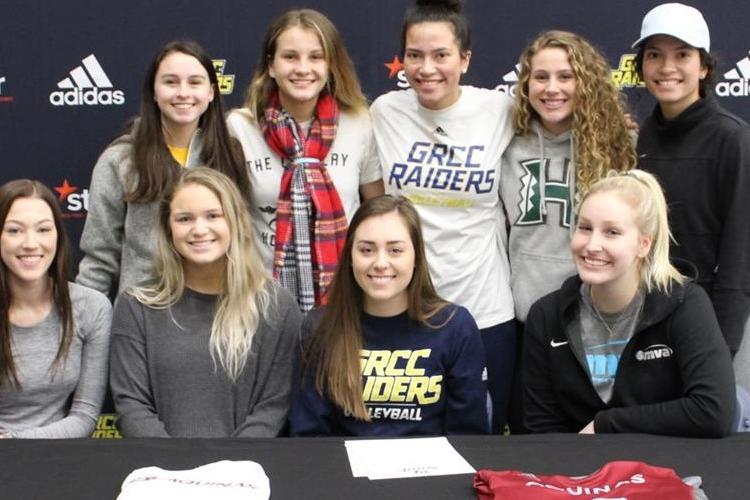 Lindsey Glaz sits at a table with her signing papers in front of her. Six young women surround her.