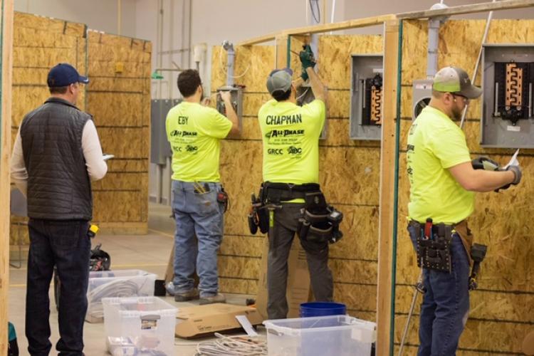 A judge, holding a clipboard, watches students perform an electrical installation at Tassell M-TEC.