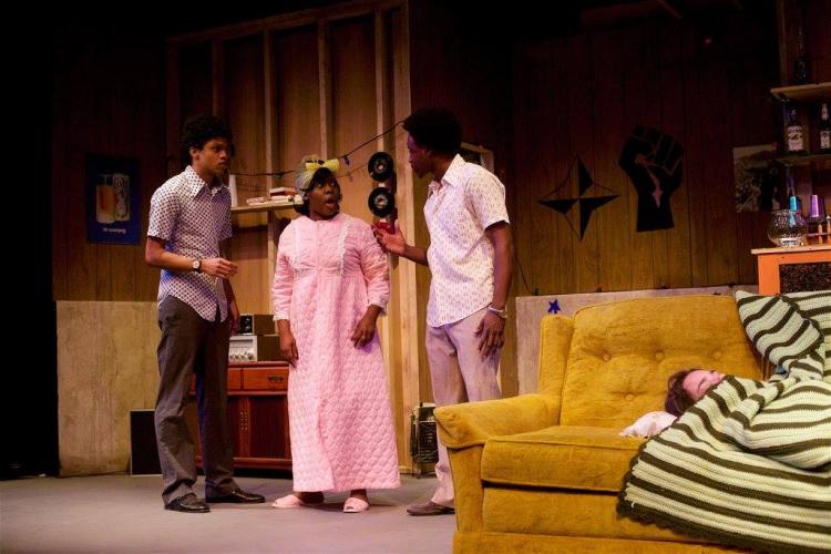 Two student actors and a student actress look at another student actress, who lies on a sofa covered by an afgan, in a scene from "Detroit '67."