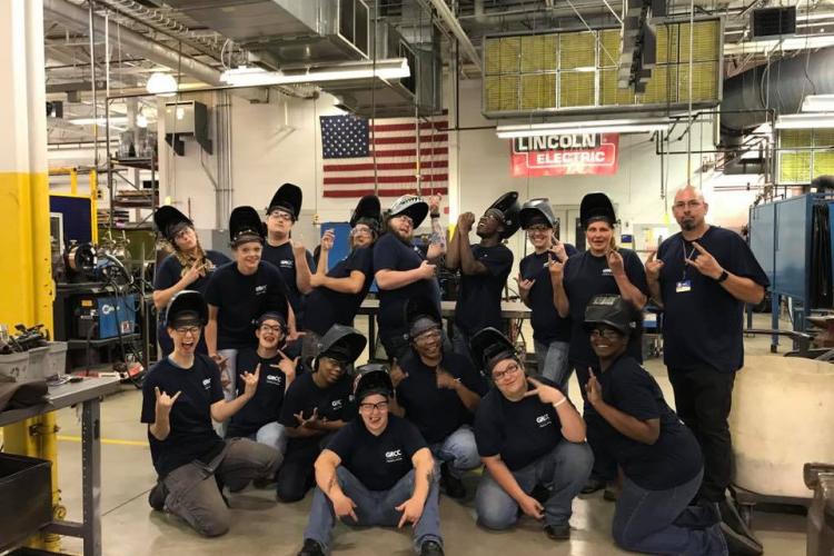 An instructor and 15 welding students, all wearing Metallica Scholars T-shirts and welding helmets, pose in Tassell M-TEC's welding facility.