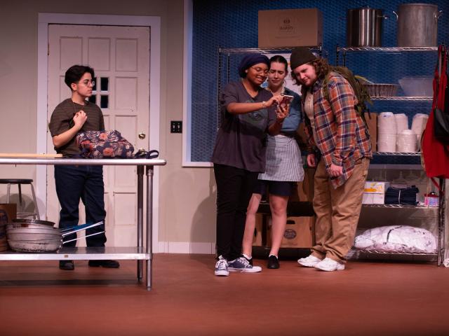 In a kitchen set on a stage, three people look at a phone while another looks on. 